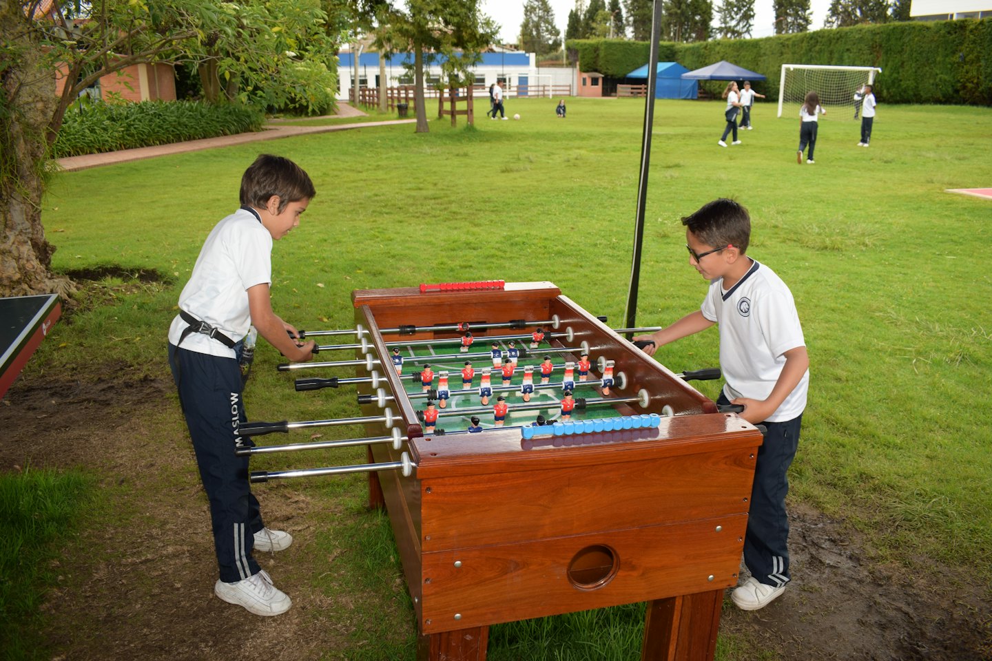 Zwei Schulkinder spielen Tischfußball an einem Holztisch im Freien.